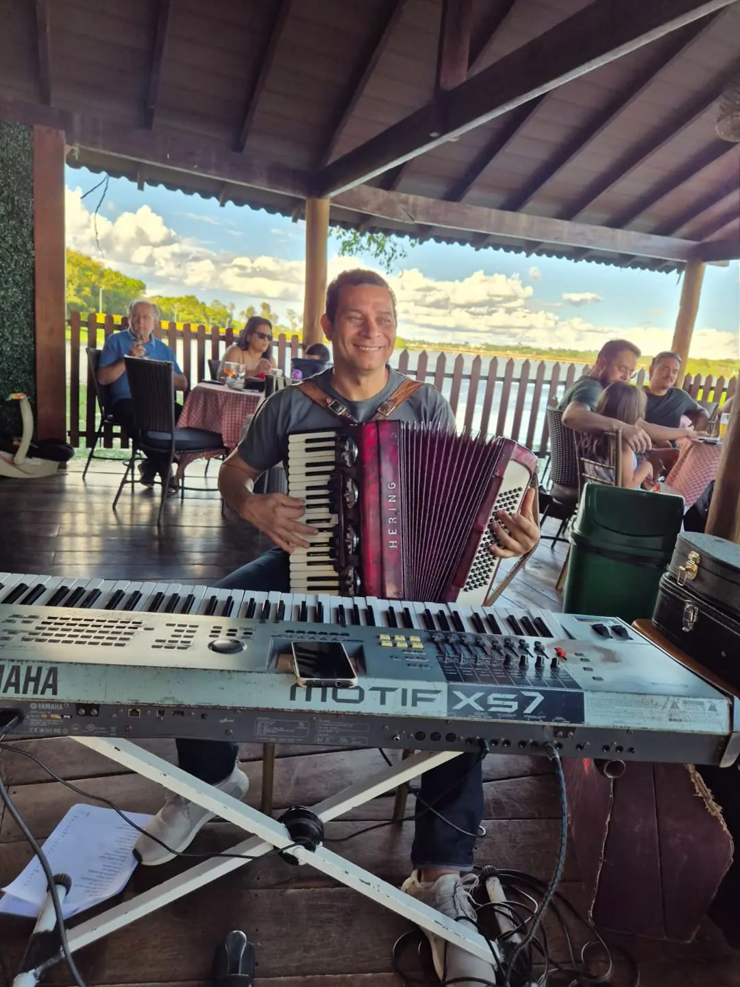 Sanfoneiro sorrindo atrás do teclado, num deque coberto à beira do lago.