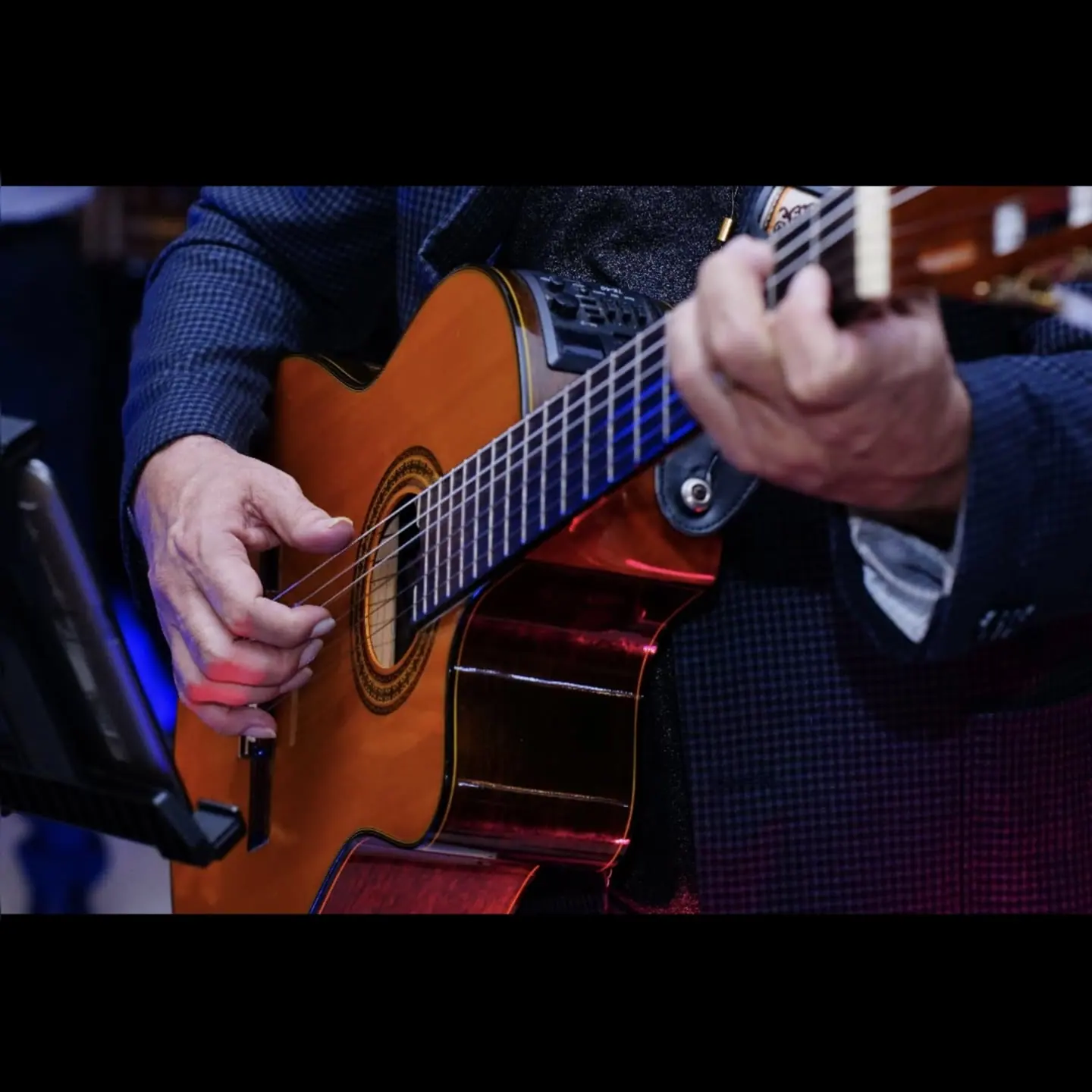 Mãos de um violonista num violão eletroacústico, sob luz azul e vermelha.